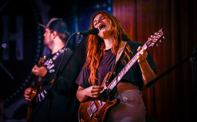 A guitarist with long red hair singing at the mic with the band behind her