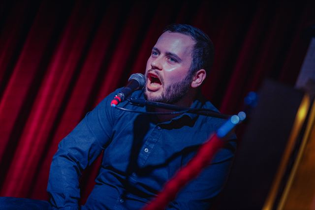 A singer in a blue shirt belting into the mic in front of a red curtain