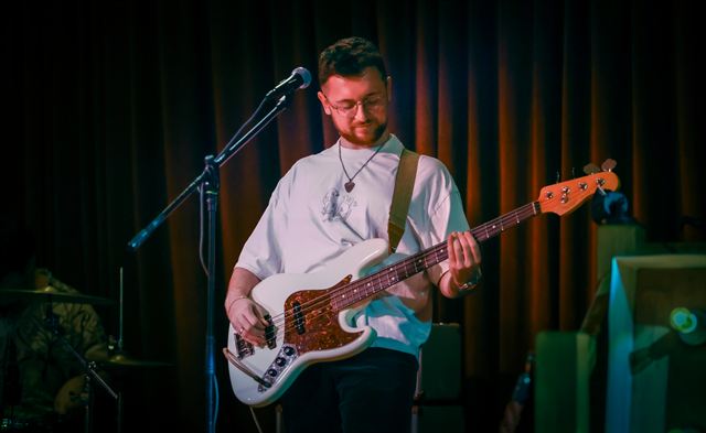 A bassist in a white t-shirt playing in front of a dark curtain