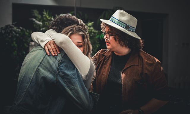 Two actors hugging in a scene while a third looks on
