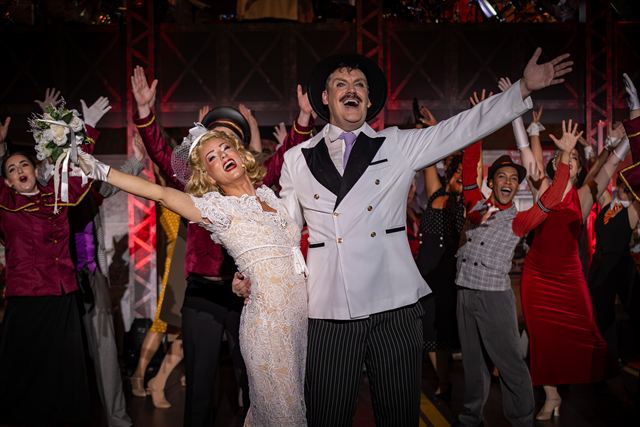 The whole cast throwing their arms up in the finale, led by a man in a white suit and a woman in a white dress