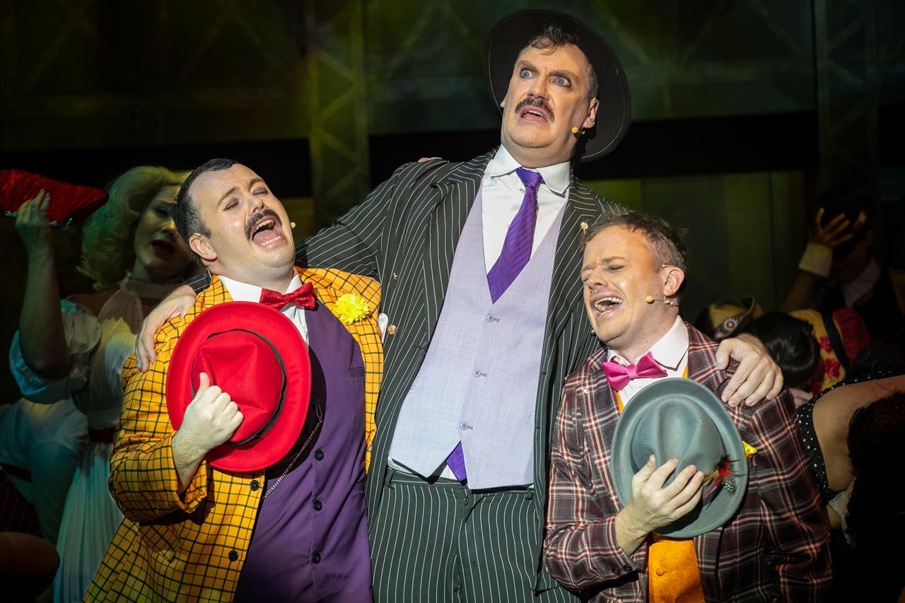 Three actors in a comic scene, one in a yellow suit holding up a red hat